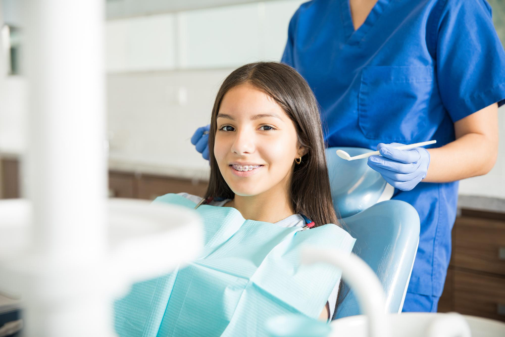 Smiling teenage girl with braces sitting in dental chair while dentist stands in clinic